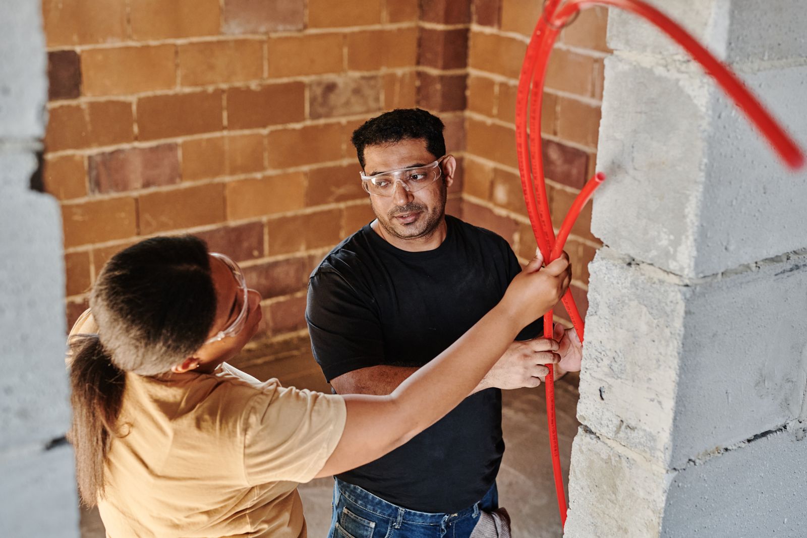 Two workers pulling red PEX water tubing through a brick wall